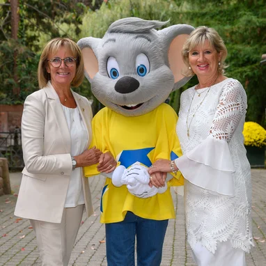 Marianne und Mauritia Mack gemeinsam mit Ed Euromaus in naturnaher Umgebung. Marianne und Mauritia Mack mit Ed Euromaus vor grüner Kulisse mit Bäumen und Blumen.